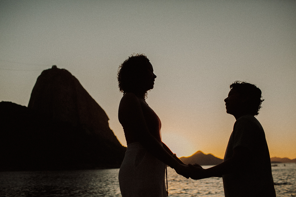 Alice e seu irmão de mãos dadas em silhueta durante ensaio de 15 anos ao pôr do sol na Praia Vermelha, Rio de Janeiro.