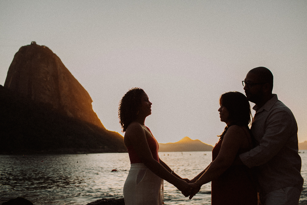 Alice recebe a bênção de seus pais durante seu ensaio fotográfico de 15 anos na Praia Vermelha, com o Pão de Açúcar ao fundo.