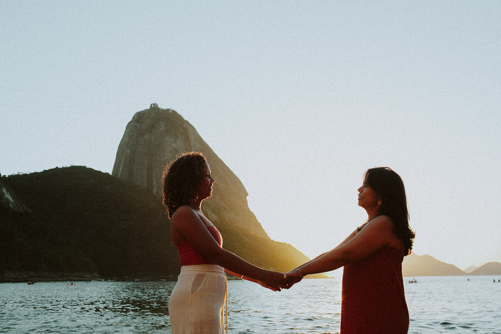 Mãe e filha de mãos dadas diante do Pão de Açúcar durante ensaio de 15 anos na Praia Vermelha, Rio de Janeiro.