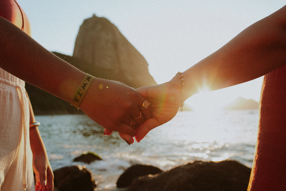 Mãos unidas ao pôr do sol durante ensaio fotográfico de 15 anos na Praia Vermelha, registrando os vínculos familiares de Alice.