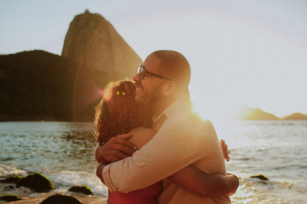 Abraço entre Alice e seu pai durante ensaio de 15 anos na Praia Vermelha, com o Pão de Açúcar e a luz do amanhecer ao fundo.