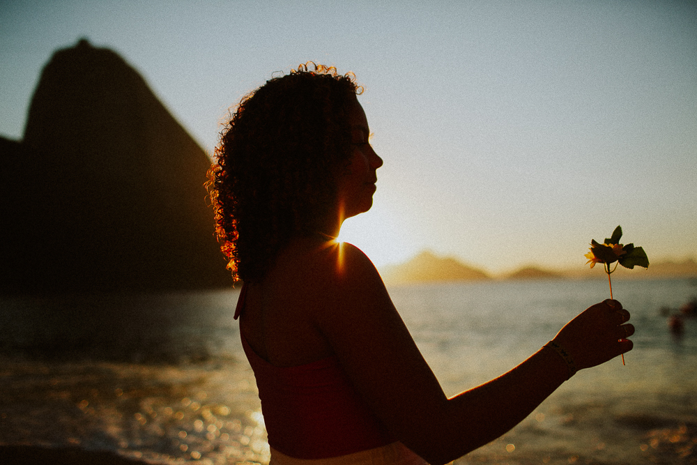Silhueta de Alice segurando uma flor ao pôr do sol durante seu ensaio fotográfico de 15 anos na Praia Vermelha.