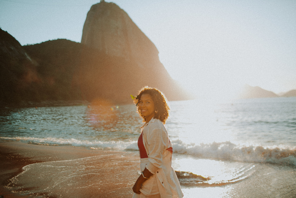 Alice caminhando pela areia durante seu ensaio de 15 anos na Praia Vermelha, com o Pão de Açúcar iluminado pelo nascer do sol