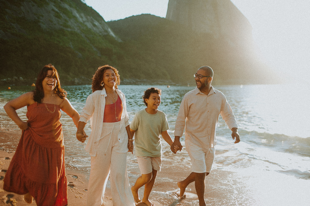 Família caminhando e sorrindo à beira-mar durante ensaio de 15 anos na Praia Vermelha, Rio de Janeiro.