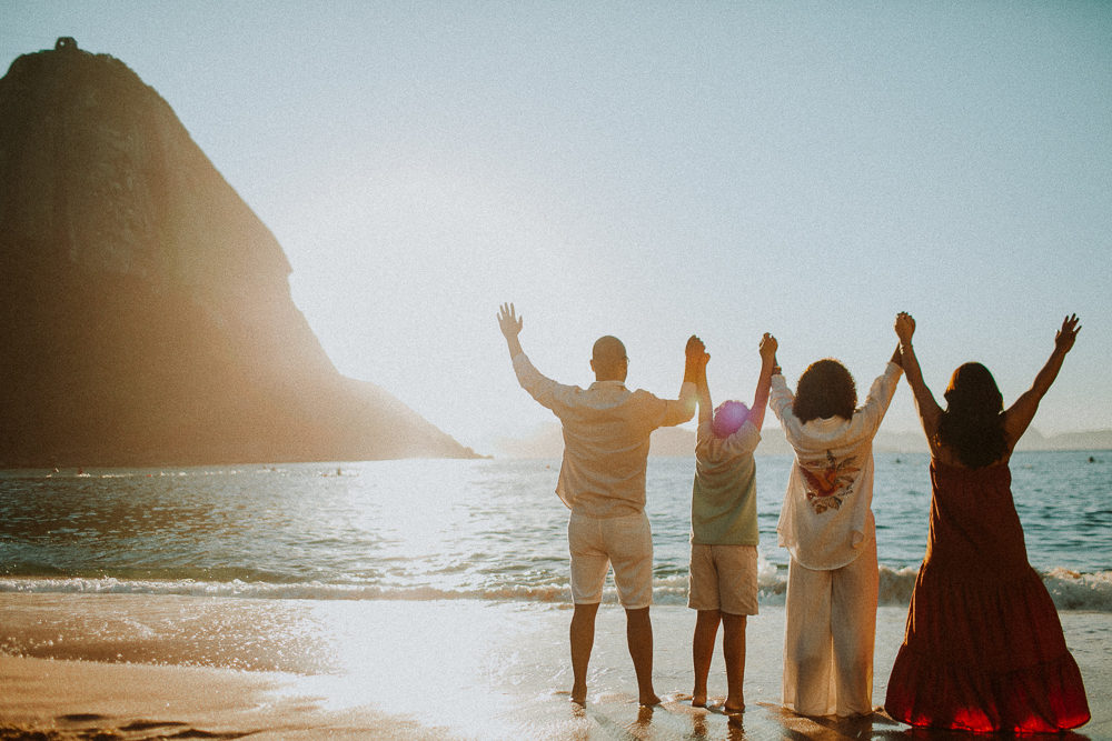 Família de Alice de mãos dadas diante do mar durante ensaio de 15 anos na Praia Vermelha, com o Pão de Açúcar ao fundo.