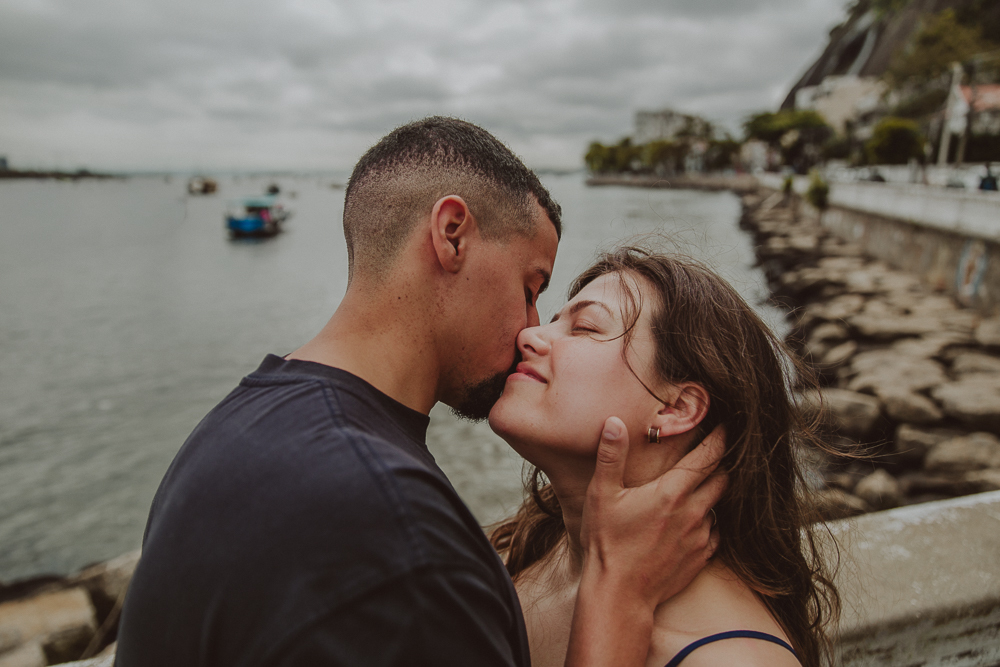 casal em um momento de carinho à beira da Baía de Guanabara durante o ensaio após o pedido de casamento na Mureta da Urca.