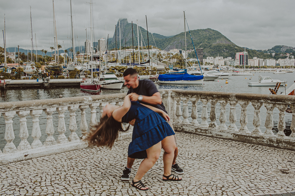 Momento espontâneo e divertido de casal durante o ensaio de casal na Urca, com a marina e as montanhas do Rio de Janeiro ao fundo.