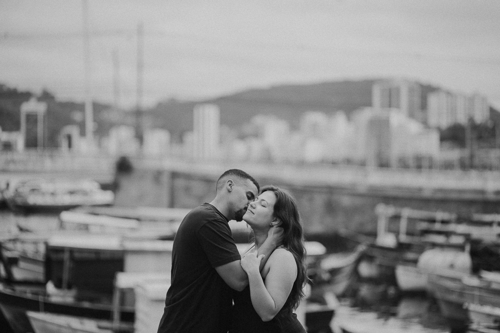casal se abraça durante o ensaio fotográfico na Urca, com barcos e a paisagem da Baía de Guanabara ao fundo.