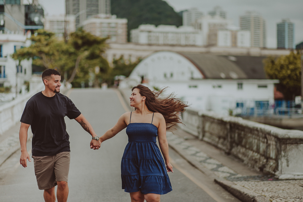 casal corre de mãos dadas pela Urca durante o ensaio de casal após o pedido de casamento, com a paisagem do Rio de Janeiro ao fundo.