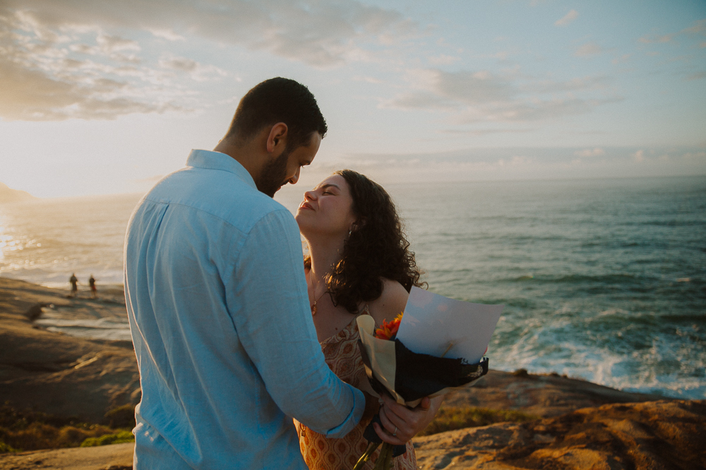 Casal se aproxima para um beijo à beira do oceano, com flores nas mãos e luz suave, na Praia do Secreto RJ.