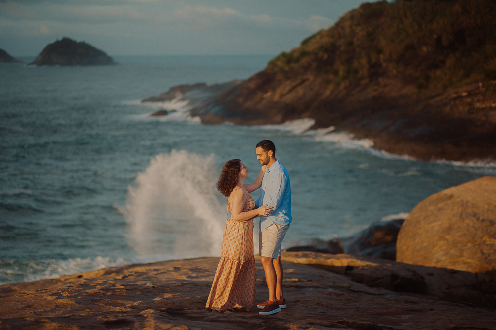 Casal permanece abraçado sobre as pedras enquanto uma onda se quebra ao fundo, na Praia do Secreto RJ.