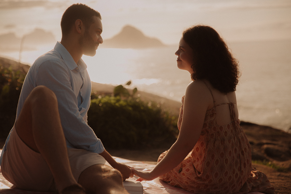 Casal sentado frente a frente, de mãos dadas, contempla a paisagem marítima ao amanhecer na Praia do Secreto RJ.