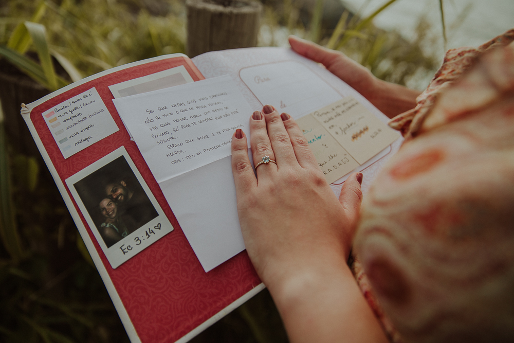 Detalhe da mão de Isabella com o anel de noivado sobre um livro aberto com mensagens e recordações (scrapbook) com a passagem Eclesiastes 3:14, após o pedido de casamento na Praia do Secreto RJ.