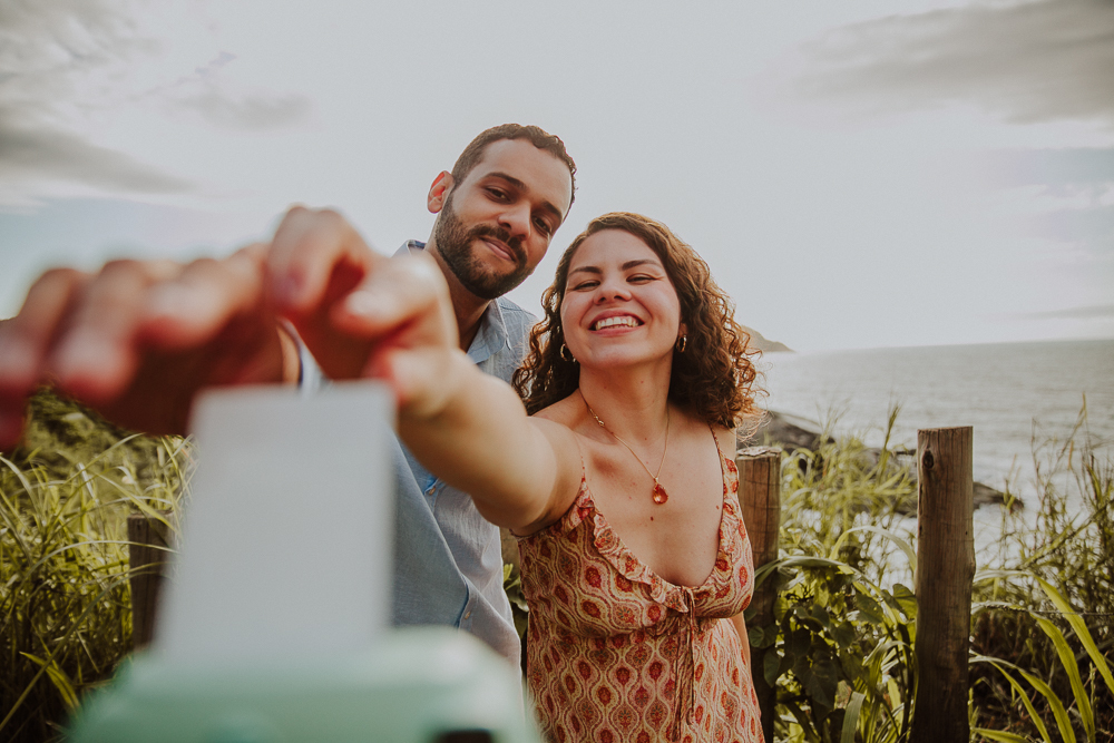 casal pega uma foto da Polaroid mostrando o anel de noivado para a câmera após o pedido de casamento, em um registro espontâneo e alegre na Praia do Secreto RJ.