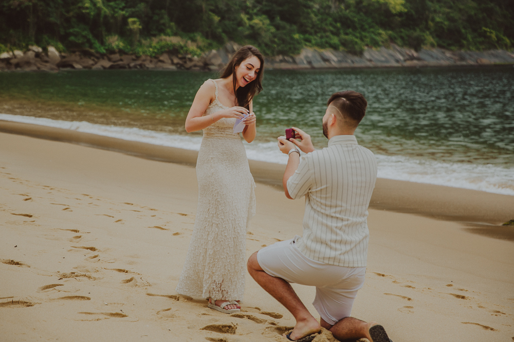 Júlia lê uma mensagem emocionada enquanto Gabriel apresenta a aliança durante o pedido de casamento na Praia Vermelha, Rio de Janeiro.