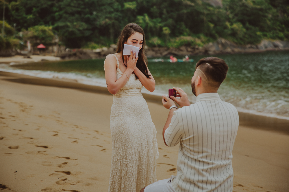 Júlia leva as mãos ao rosto, surpresa com o pedido de casamento de Gabriel, na Praia Vermelha, Urca, Rio de Janeiro.