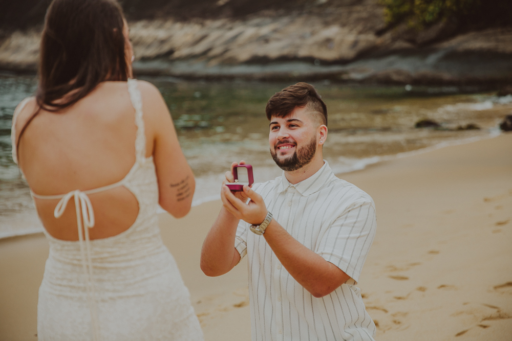 Gabriel, ajoelhado com a caixinha da aliança aberta, olha para Júlia durante o pedido de casamento na Praia Vermelha, RJ.