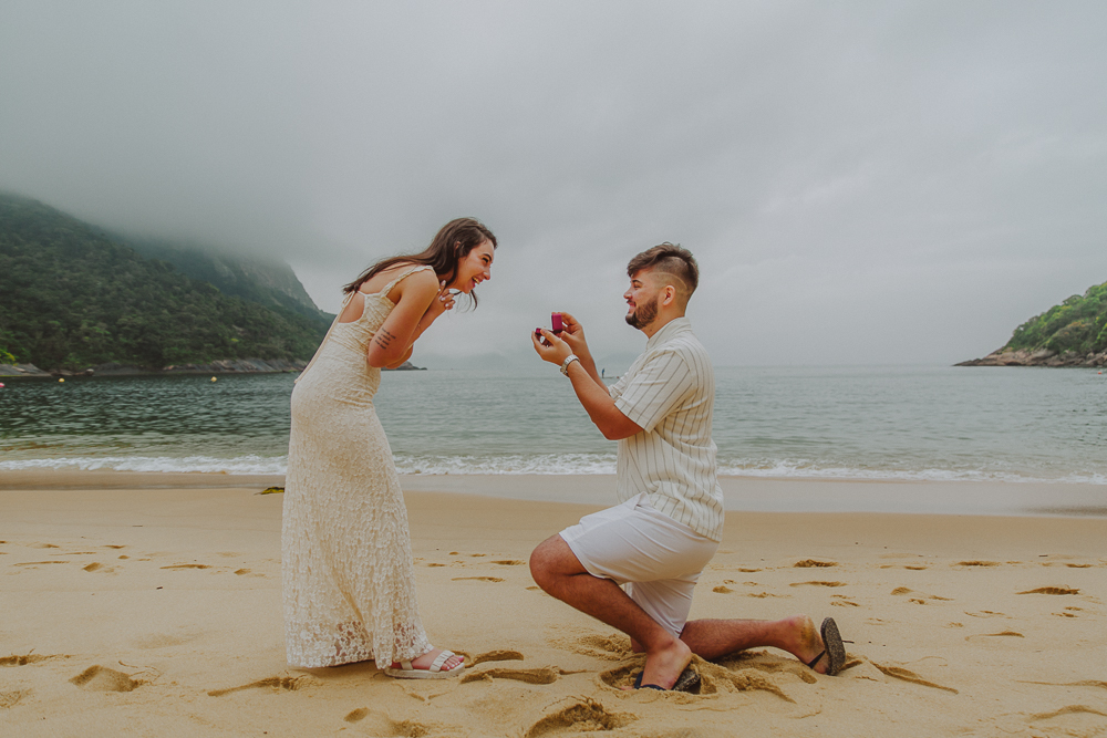 Júlia se inclina emocionada para Gabriel durante o pedido de casamento surpresa, com o mar e as montanhas da Praia Vermelha ao fundo.