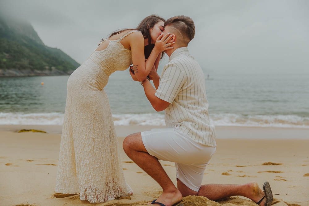 Gabriel e Júlia se beijam após o pedido de casamento, com o mar e o céu nublado da Praia Vermelha, no Rio de Janeiro, ao fundo.