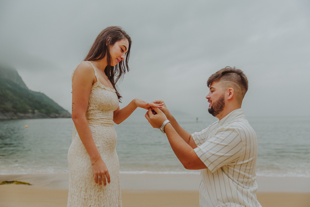 Gabriel coloca o anel de noivado no dedo de Júlia durante o pedido de casamento na Praia Vermelha, Urca, Rio de Janeiro.