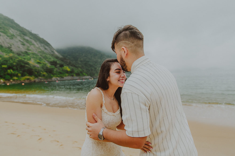 Gabriel envolve Júlia em um abraço carinhoso durante o ensaio de casal após o pedido de casamento na Praia Vermelha, RJ.
