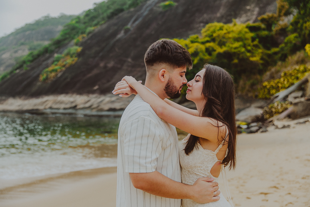 Gabriel e Júlia se abraçam diante da vegetação e das pedras da Praia Vermelha durante o ensaio fotográfico de noivado no Rio de Janeiro.