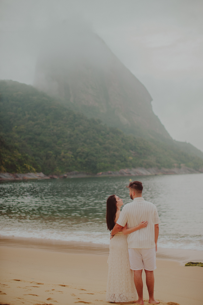 Gabriel e Júlia contemplam o mar abraçados, de costas para a câmera, durante o ensaio após o pedido de casamento na Praia Vermelha.