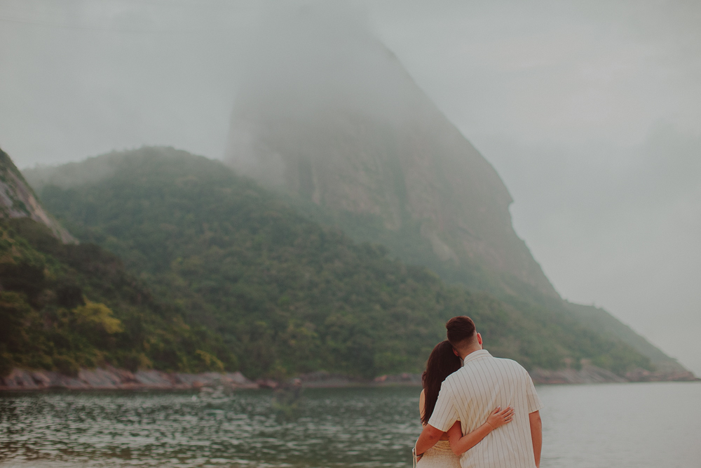 Gabriel e Júlia abraçados diante do mar e das montanhas cobertas por nuvens, durante o ensaio fotográfico na Praia Vermelha, Rio de Janeiro.
