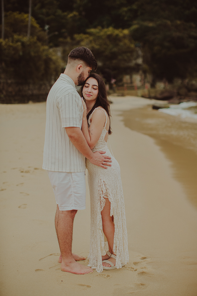 Júlia sorri de olhos fechados enquanto Gabriel a abraça pela cintura, em um retrato espontâneo na Praia Vermelha, Urca.