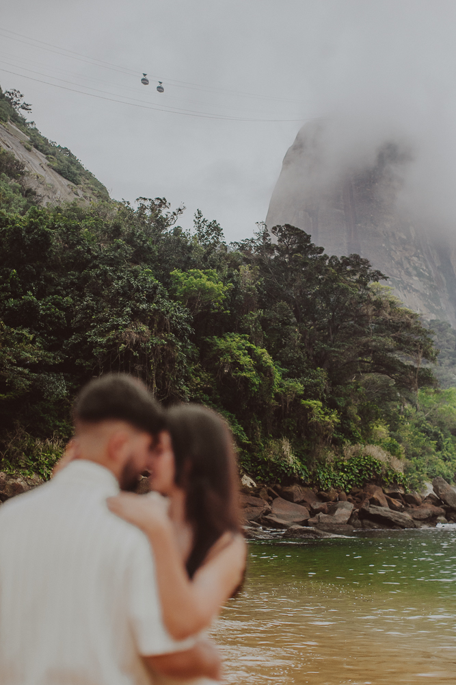 Retrato de Gabriel e Júlia com a montanha parcialmente encoberta por nuvens ao fundo, durante o ensaio de noivado na Praia Vermelha. Fotografia desfocada é arte.