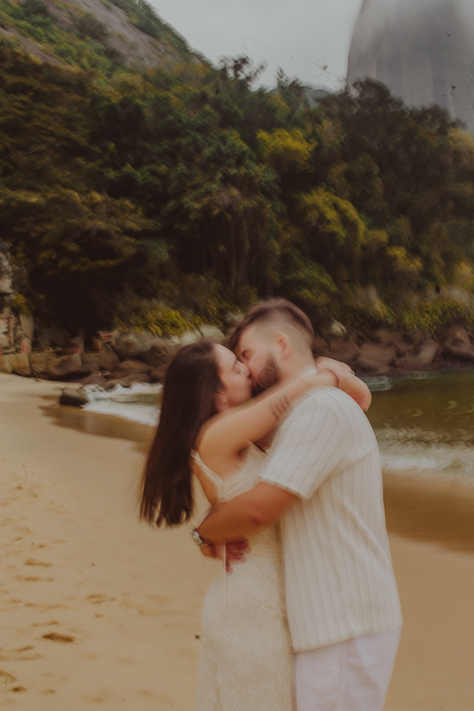Gabriel e Júlia se beijam abraçados perto das pedras e da vegetação costeira da Praia Vermelha, Urca, Rio de Janeiro. Fotografia desfocada é arte. Baixa velocidade obturador camera fotografica