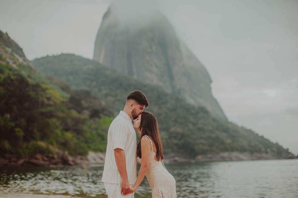 Gabriel e Júlia de mãos dadas diante do mar, com a montanha coberta por nuvens ao fundo, em ensaio de noivado na Praia Vermelha.