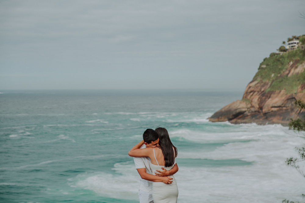 Diego y María se abrazan frente al océano después de su propuesta de matrimonio en el Mirante da Joatinga, Río de Janeiro.