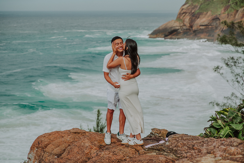 Pareja colombiana abrazada sobre las rocas, con las olas del mar de Joatinga como escenario de su pedida de mano en Brasil.
