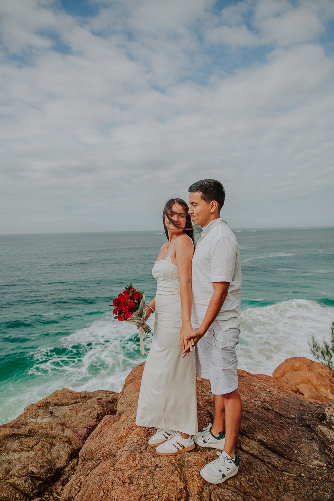 María sostiene un ramo de rosas rojas junto a Diego durante su sesión de fotos de compromiso frente al mar en Joatinga.