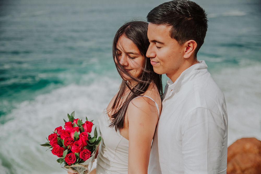 Retrato romántico de Diego y María con un ramo de rosas rojas y las olas del océano al fondo, en Río de Janeiro, Brasil.