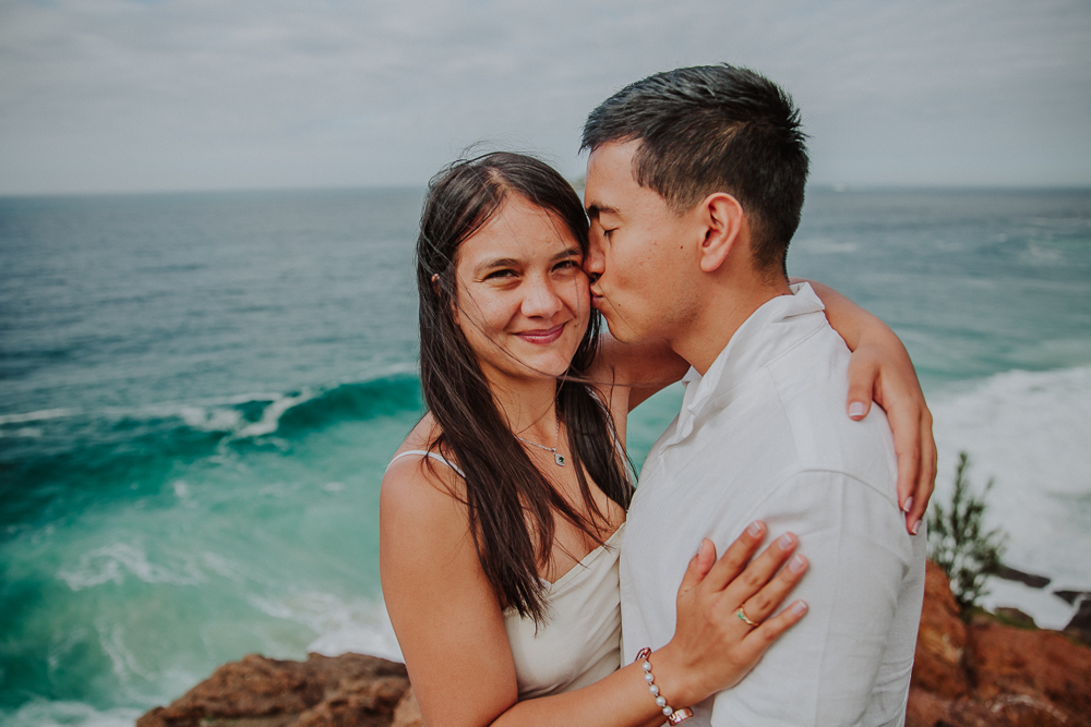 Diego besa la mejilla de María mientras ella sonríe frente al mar, después de su propuesta de matrimonio en Río de Janeiro.