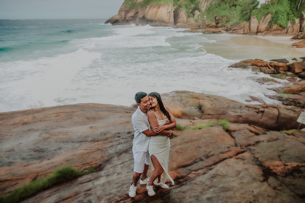 Diego y María se abrazan sobre las rocas con la playa y las olas al fondo, en el Mirante da Joatinga, Brasil.