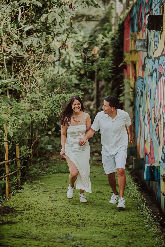 Diego y María caminan de la mano por un sendero rodeado de vegetación, junto a un muro con arte urbano en Joatinga.