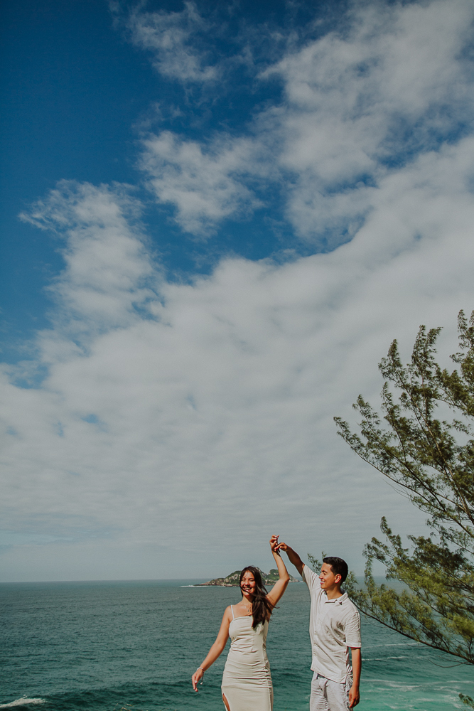 Diego y María bailan frente al mar, bajo un cielo azul con nubes blancas y entre las ramas de los árboles, durante su sesión de compromiso en el Mirante da Joatinga, Río de Janeiro, Brasil.
