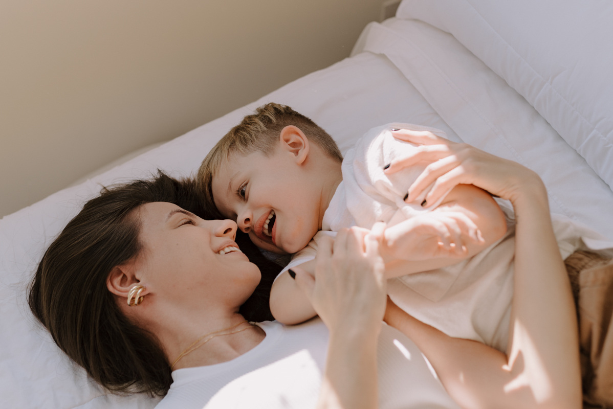 Mãe deitada na cama, brincando e sorrindo com seu filho pequeno, capturando a essência de momentos carinhosos e espontâneos no dia a dia.