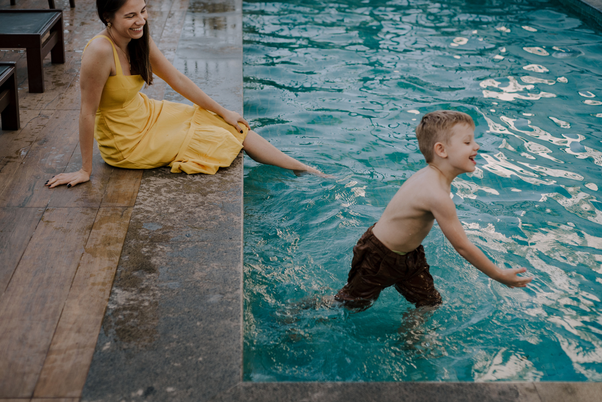 Mãe sentada ao lado da piscina, observando o filho brincar na água, refletindo a beleza dos pequenos momentos cotidianos.
