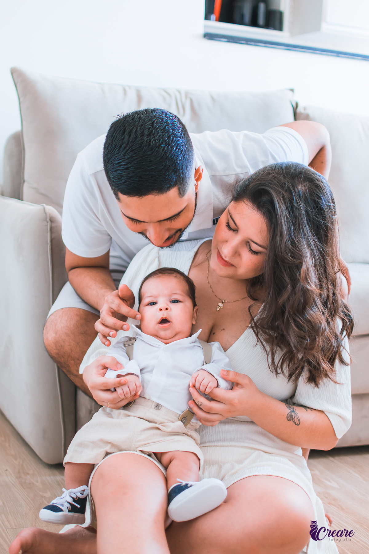 Ensaio newborn lifestyle, ensaio família. A imagem contém: um homem jovem com roupa branca, uma mulher jovem com roupa branca, um bebê com roupa branca e bermuda bege com suspensório. fotógrafo Mauá, ensaio lifestyle. 