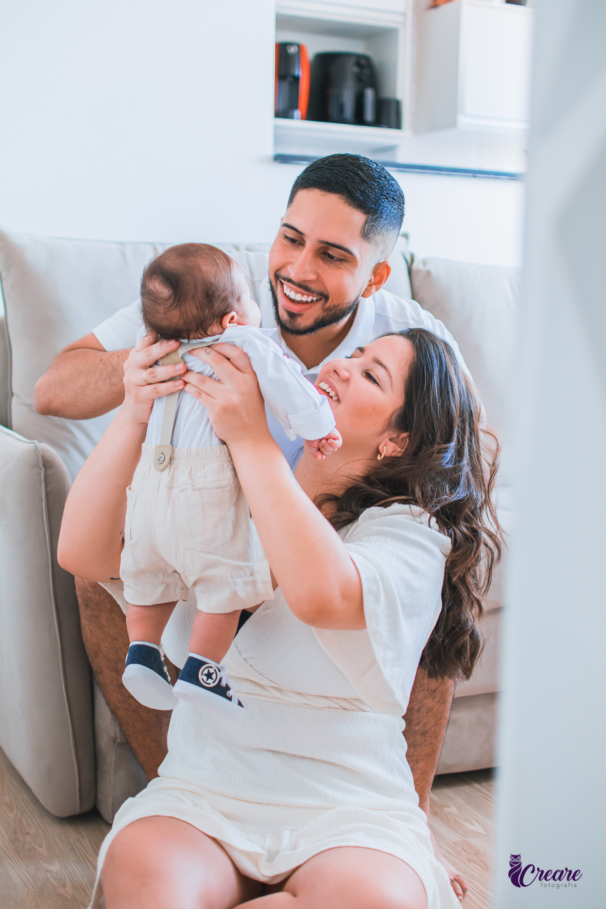 Ensaio newborn lifestyle, ensaio família. A imagem contém: um homem jovem com roupa branca, uma mulher jovem com roupa branca, um bebê com roupa branca e bermuda bege com suspensório. fotógrafo Mauá, ensaio lifestyle. 