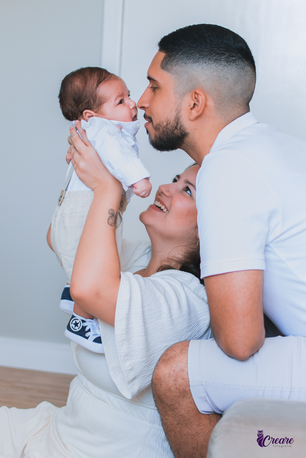 Ensaio newborn lifestyle, ensaio família. A imagem contém: um homem jovem com roupa branca, uma mulher jovem com roupa branca, um bebê com roupa branca e bermuda bege com suspensório. fotógrafo Mauá, ensaio lifestyle. 