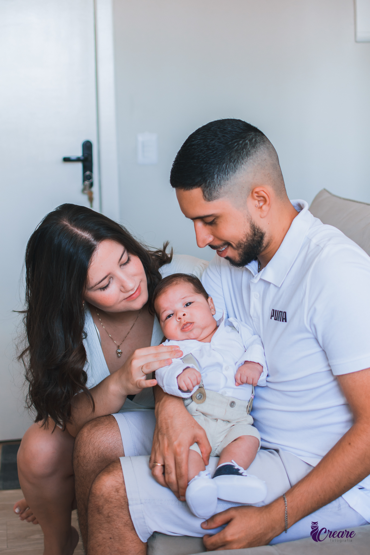 Ensaio newborn lifestyle, ensaio família. A imagem contém: um homem jovem com roupa branca, uma mulher jovem com roupa branca, um bebê com roupa branca e bermuda bege com suspensório. fotógrafo Mauá, ensaio lifestyle. 