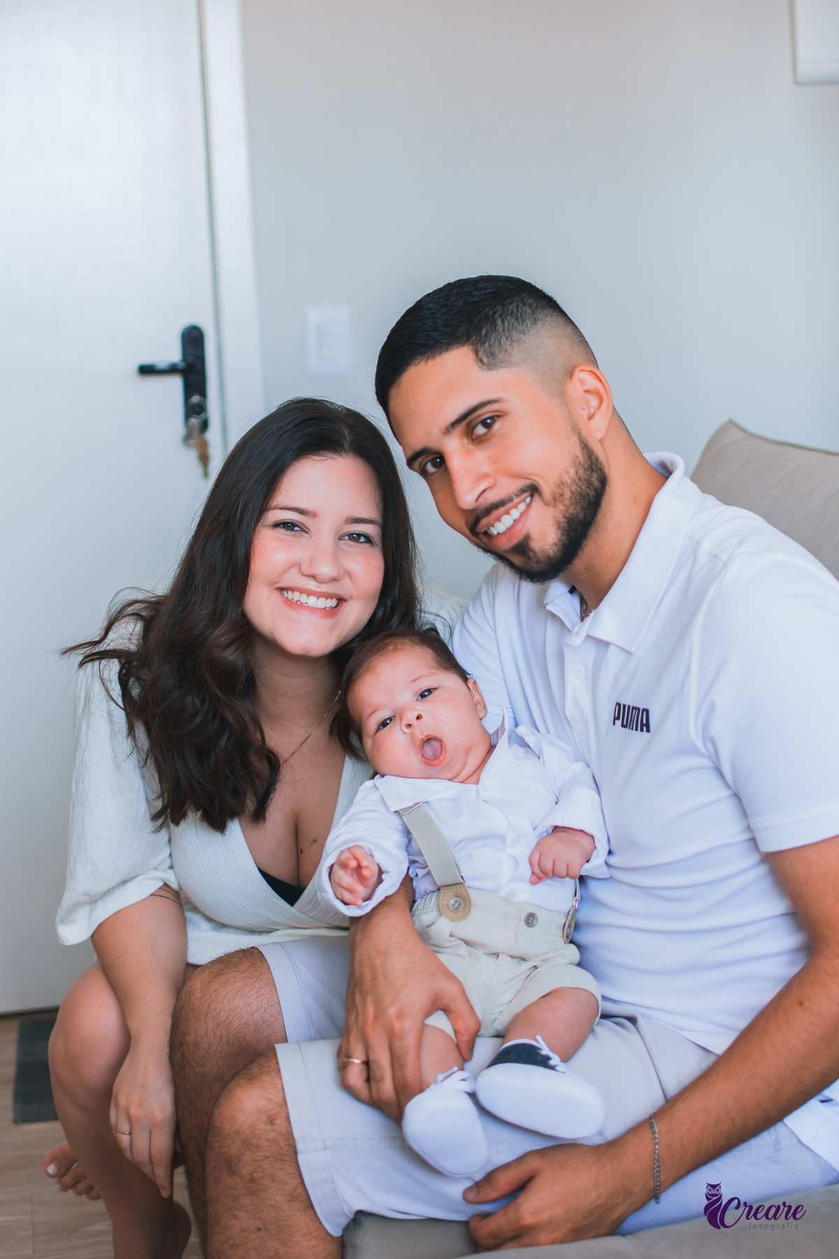 Ensaio newborn lifestyle, ensaio família. A imagem contém: um homem jovem com roupa branca, uma mulher jovem com roupa branca, um bebê com roupa branca e bermuda bege com suspensório. fotógrafo Mauá, ensaio lifestyle. 