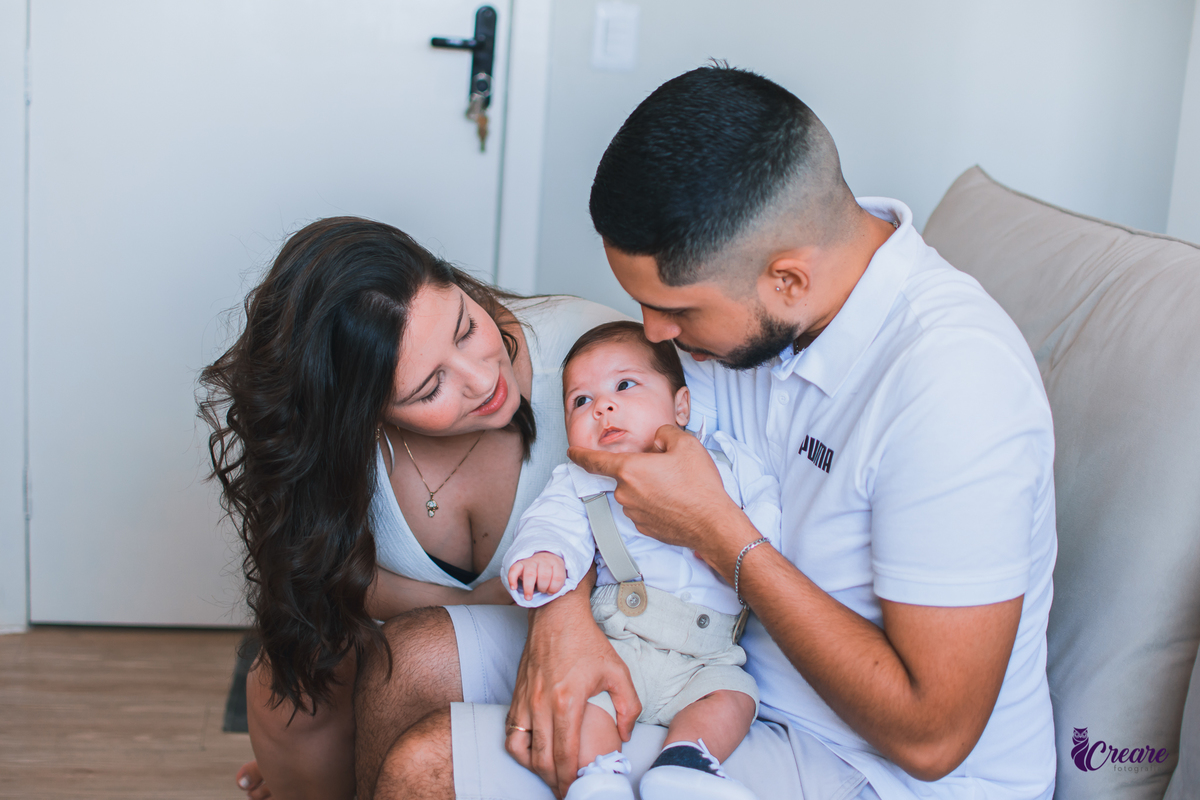 Ensaio newborn lifestyle, ensaio família. A imagem contém: um homem jovem com roupa branca, uma mulher jovem com roupa branca, um bebê com roupa branca e bermuda bege com suspensório. fotógrafo Mauá, ensaio lifestyle. 