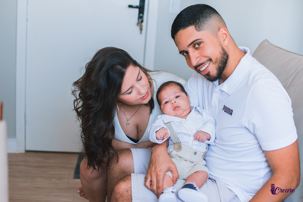 Ensaio newborn lifestyle, ensaio família. A imagem contém: um homem jovem com roupa branca, uma mulher jovem com roupa branca, um bebê com roupa branca e bermuda bege com suspensório. fotógrafo Mauá, ensaio lifestyle. 