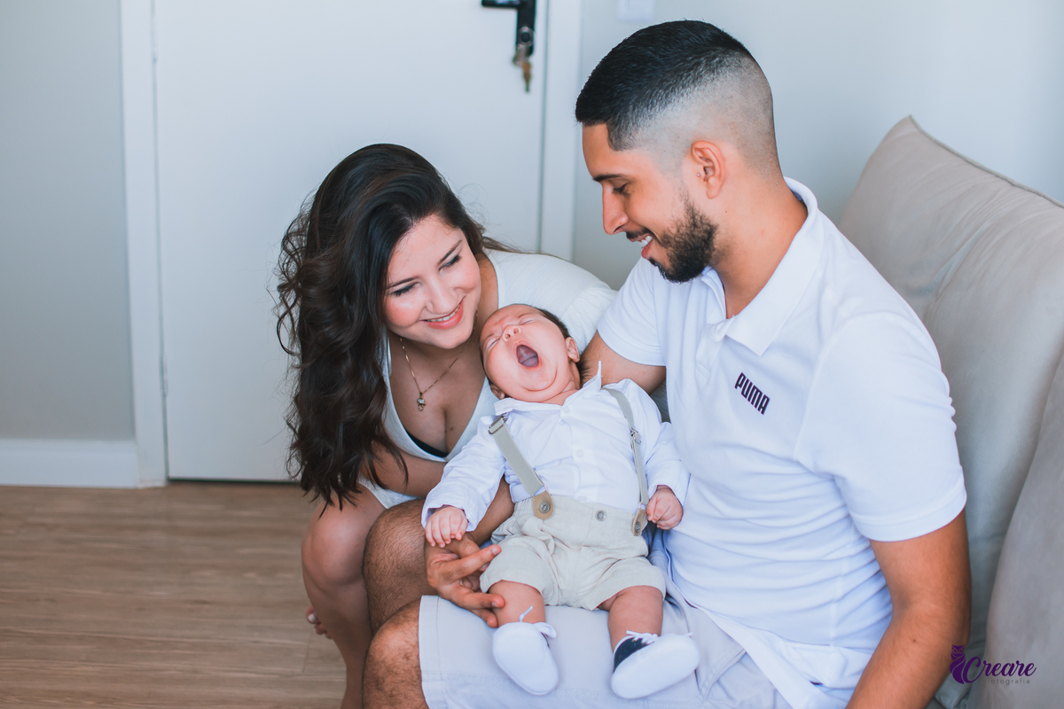 Ensaio newborn lifestyle, ensaio família. A imagem contém: um homem jovem com roupa branca, uma mulher jovem com roupa branca, um bebê com roupa branca e bermuda bege com suspensório. fotógrafo Mauá, ensaio lifestyle. 